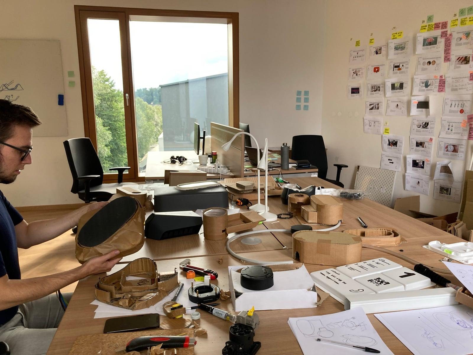 Designer handling cardboard and wooden form models at a cluttered studio desk