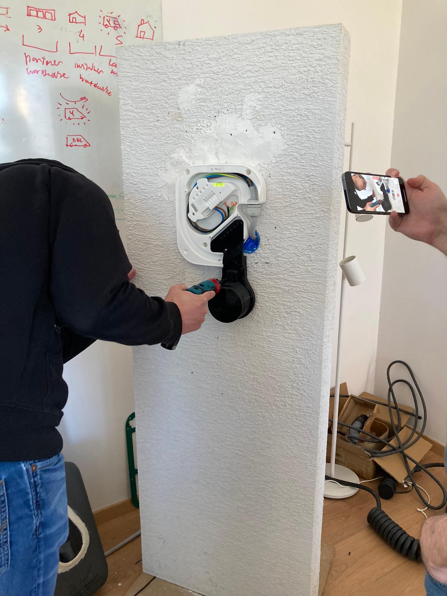 Testing a charger prototype mounted on a mock-up wall while a colleague records on a phone