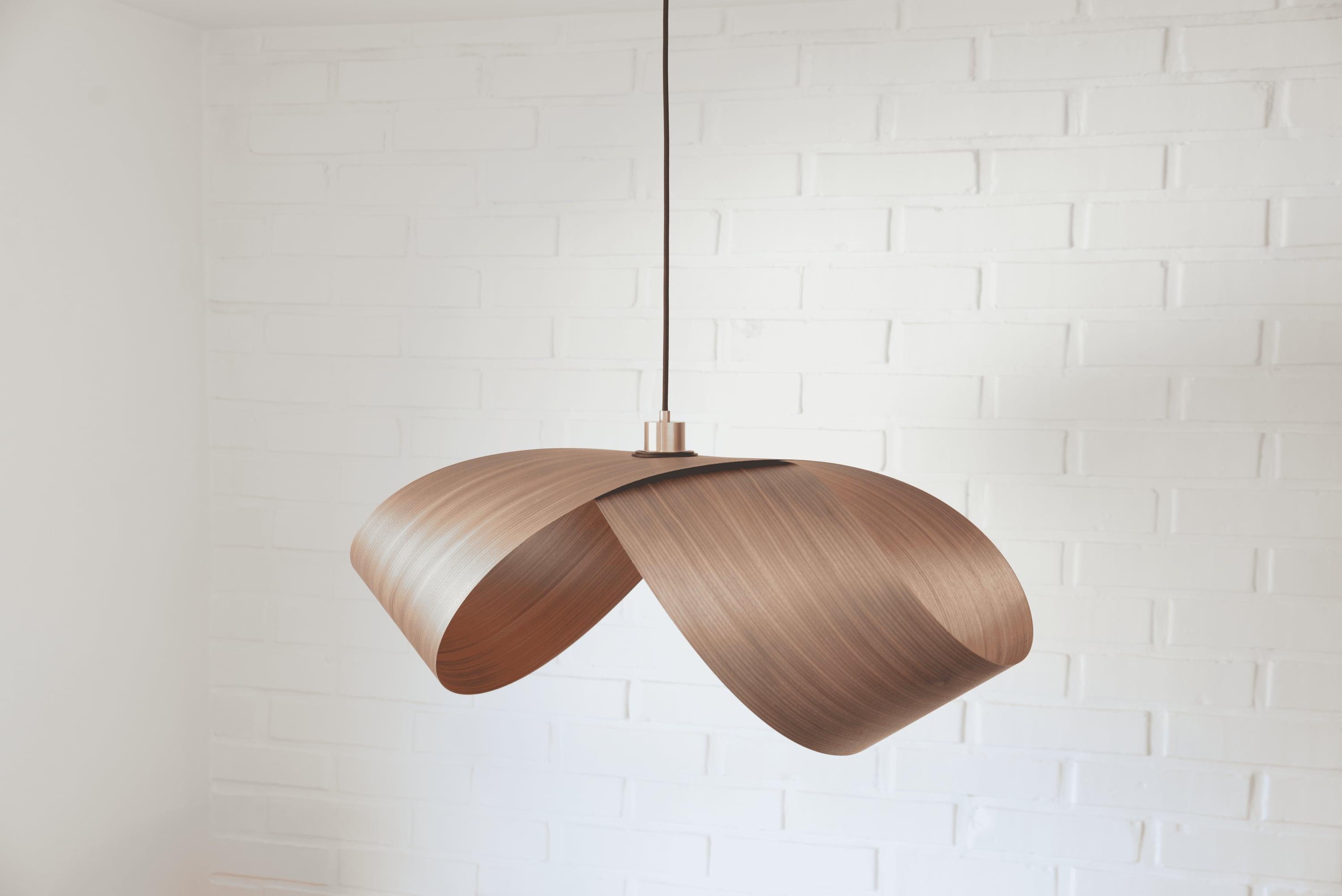 Foldia pendant lamp with a folded walnut wood-veneer shade hanging in front of a white brick wall