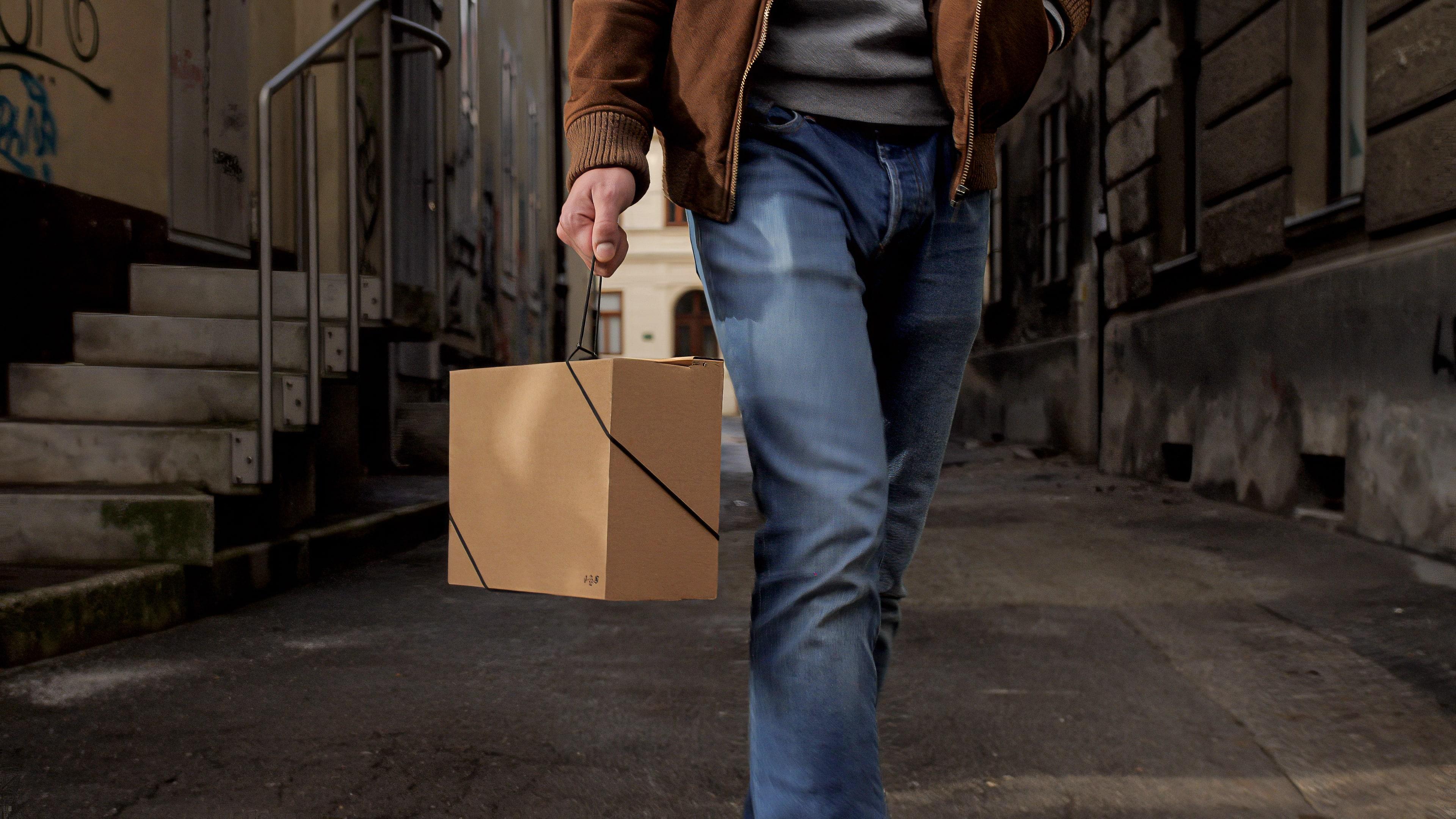 Person walking down a city street carrying a cardboard box by its Bagless Cord handle