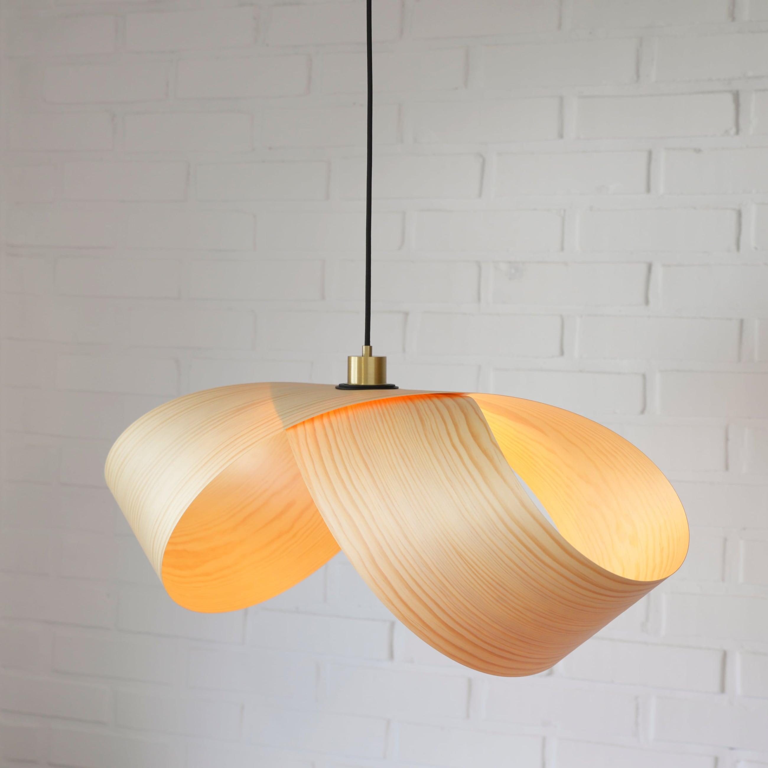 Foldia lamp in pine veneer, open and lit from inside, in front of a white brick wall