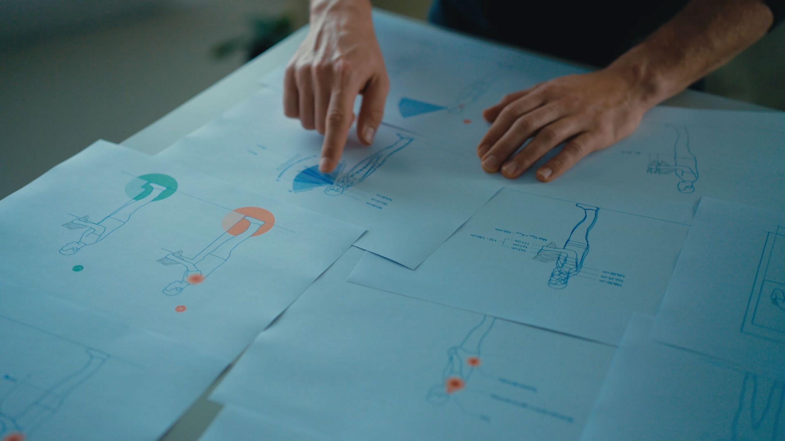 Two hands over spread sheets of blue ergonomic drawings, one finger pointing at a viewing-angle diagram