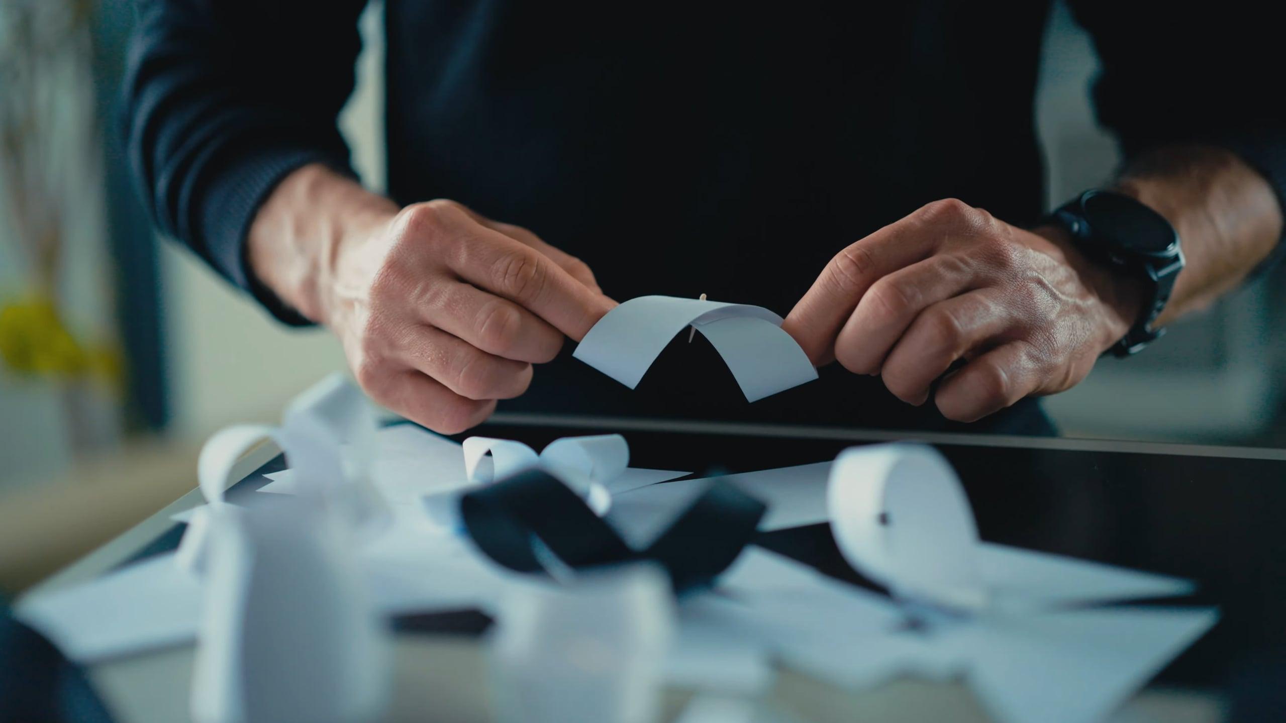 Hands bending a strip of white paper into a curve above a desk covered with paper mockups
