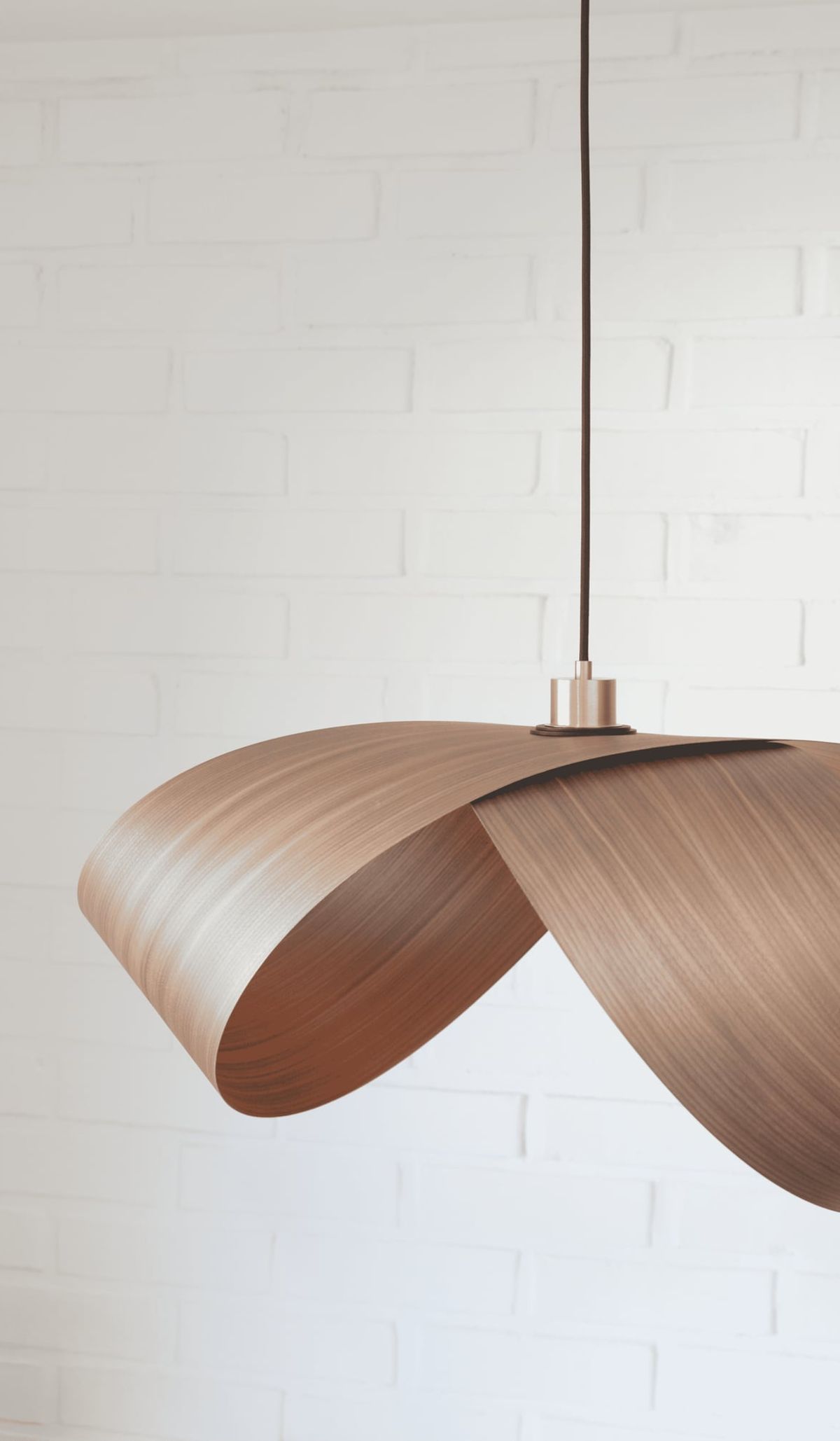 Foldia pendant lamp with a folded walnut wood-veneer shade hanging in front of a white brick wall